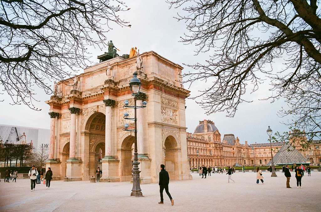 An archway near the Louvre frame by trees, with a glass pyramid visible in the background