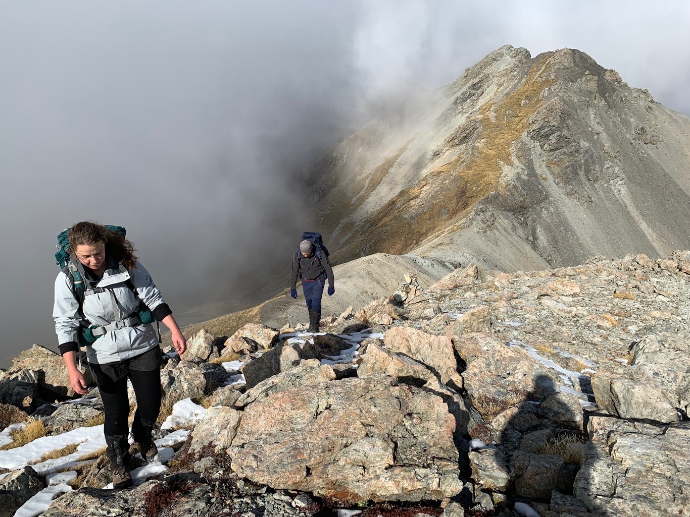 Two people with tramping packs walking along a ridge with grey cloud, rocks and short grassy slopes