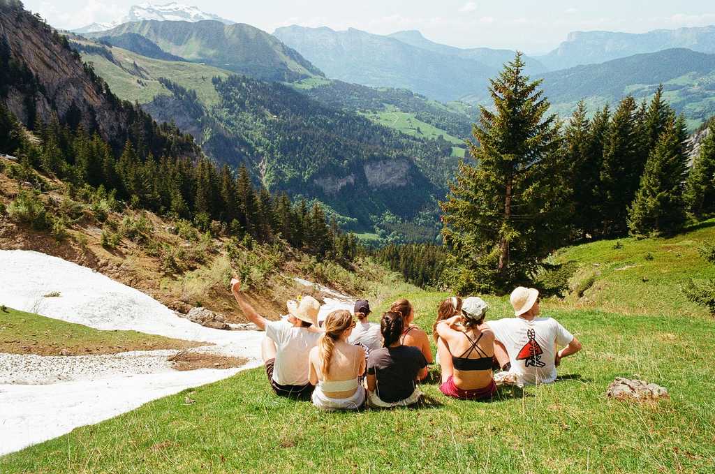 The backs of eight people sitting on grass looking at an alpine view of trees, snow and mountains