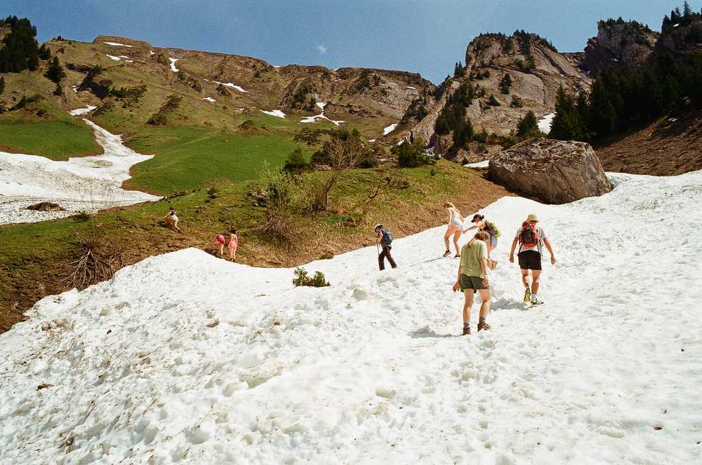 A group of people in a S-shaped line crossing a patch of snow onto a grassy alpine slope