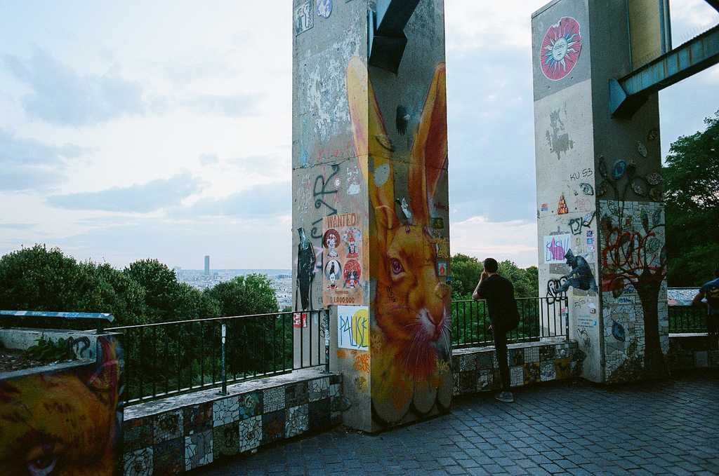 Graffiti covered concrete posts in the foreground with a man looking contemplatively out over the view of Paris from the Parc de Belleville, with the Tour Montsouris visible in the distance