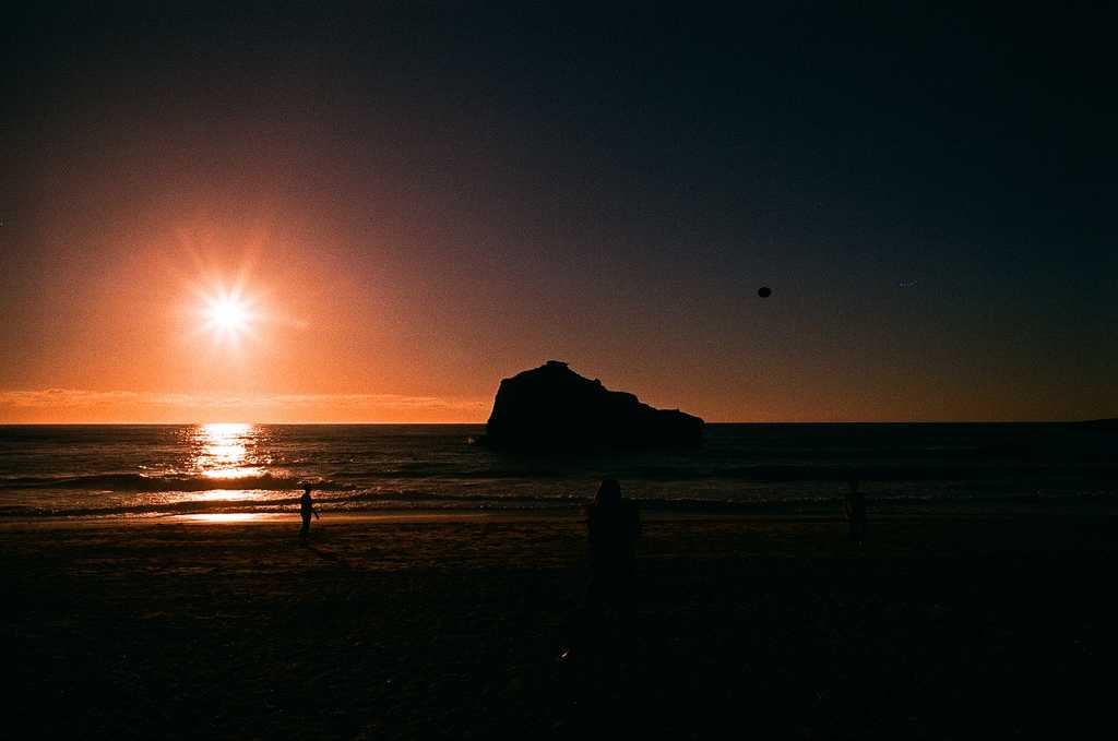 A sunset beach scene with low reddish light, rocky outcrops in the sea and silhouettes playing with a ball on the beach.