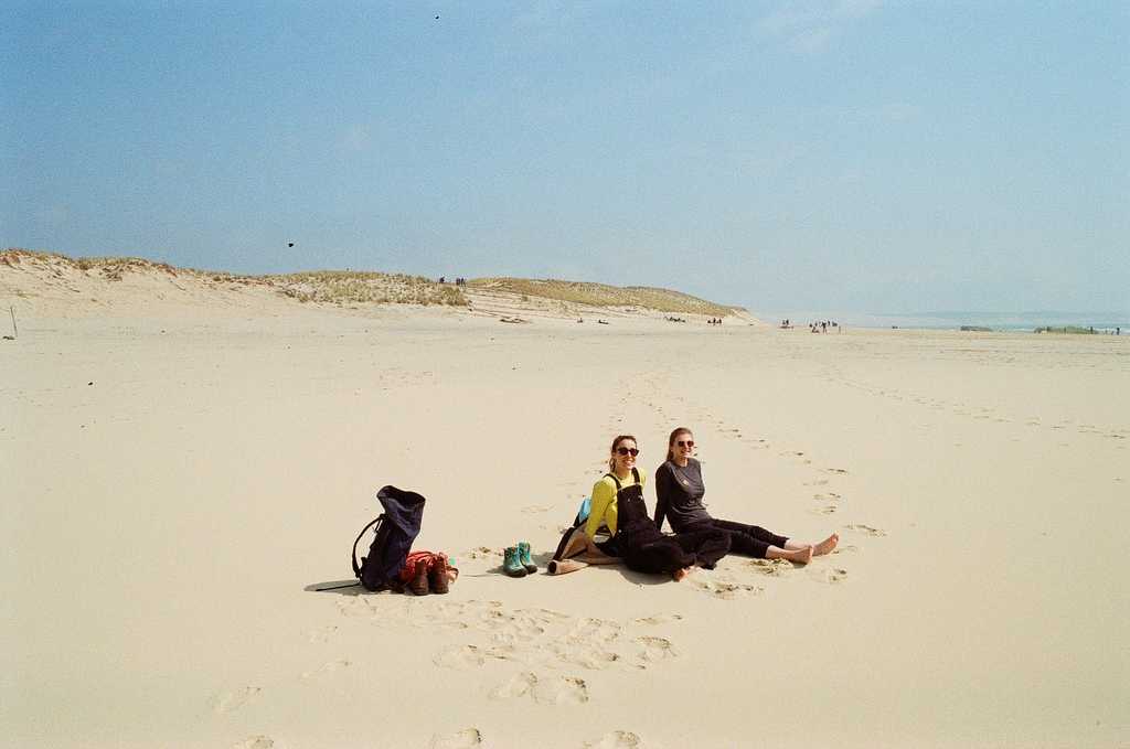 Two women sitting on a wide sandy beach, with sand dunes in the distant