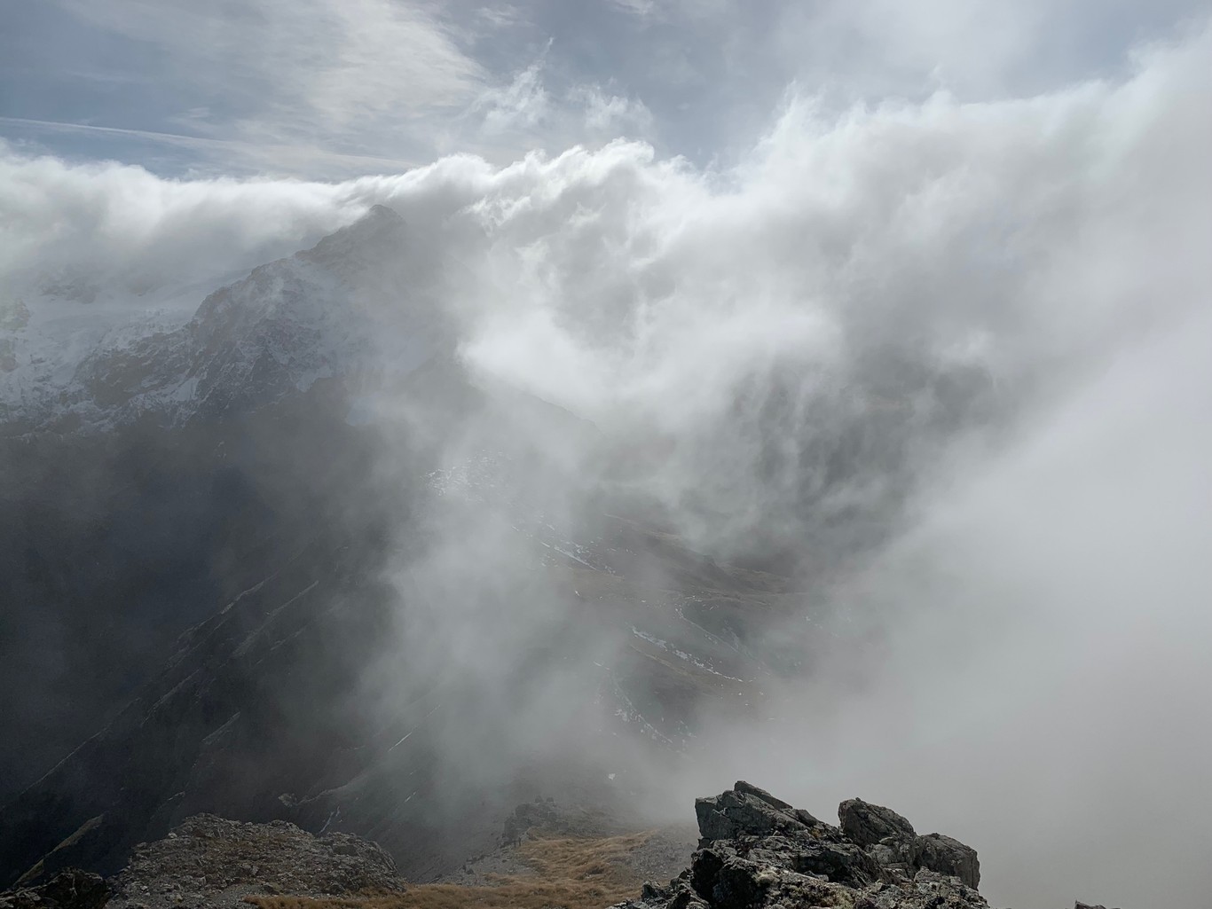 A view of an alpine ridge shrouded in clouds, with rocky, snow-covered peaks in the distance